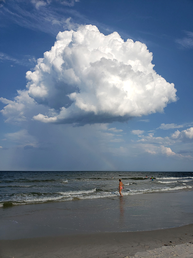 a view from a beach out to the ocean, featuring a large raincloud, rainbow, and a woman in a bikini returning to the water after the storm