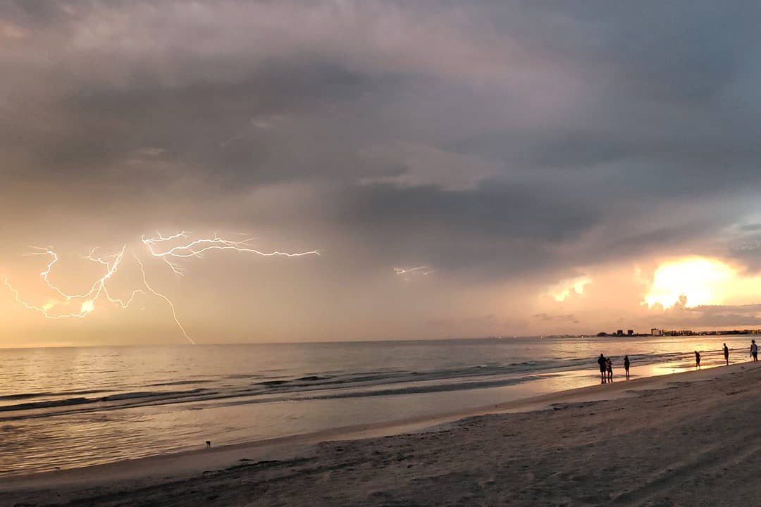 a photo of a beach at sunset, with lightning in the background